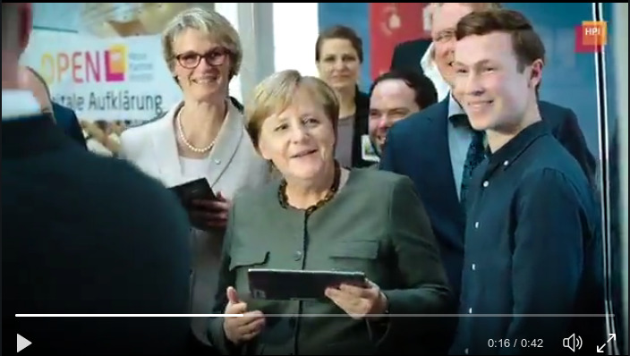 TThe image shows the German chancellor holding a tablet PC, surrounded by colleagues and others