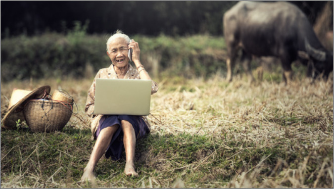 The image shows an elderly lady sitting in a field with a laptop on her knees and a smartphone near her ear. In the background a water buffalo.