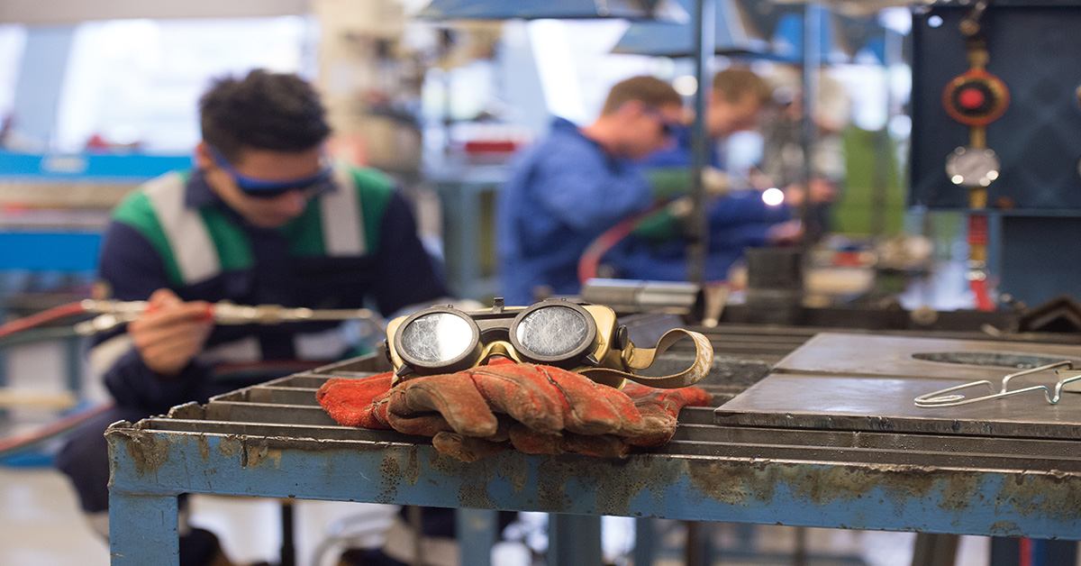 Safety goggles and gloves on top of machine parts in factory, with workers visible in background