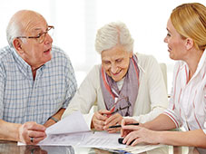 Coppia di anziani seduta a un tavolo che discute alcuni documenti con un'addetta all'assistenza