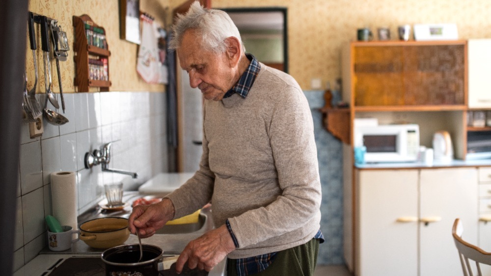 Elderly man is standing in a kitchen and cooking something small in one pot