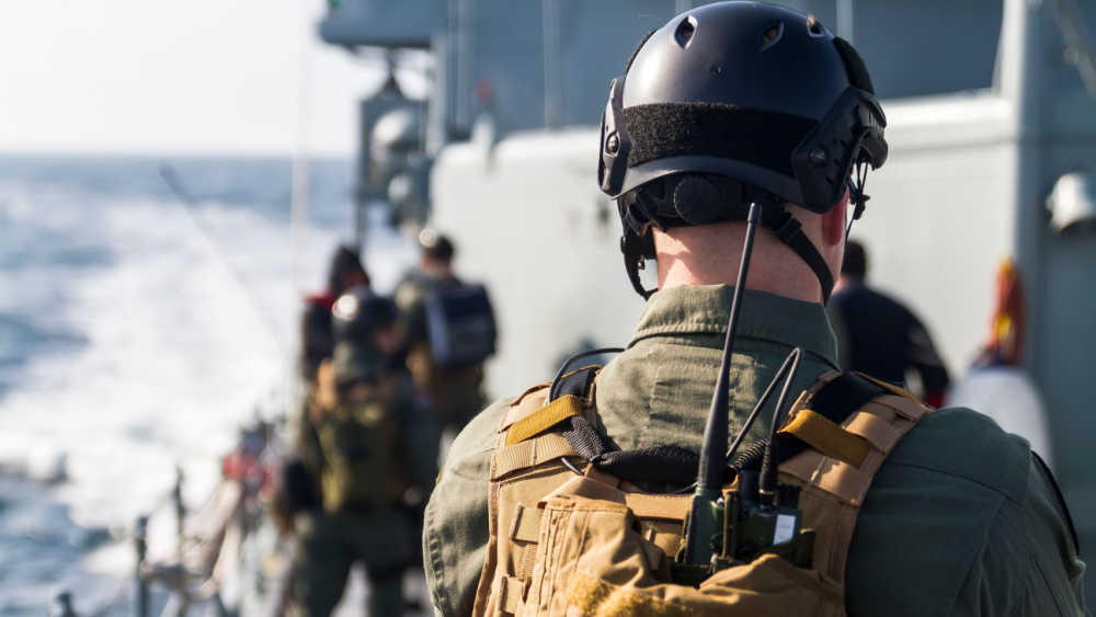 A person wearing what appears to be a uniform, with a helmet and a communication device stands with his back to the camera on the boat deck with other uniformed people.