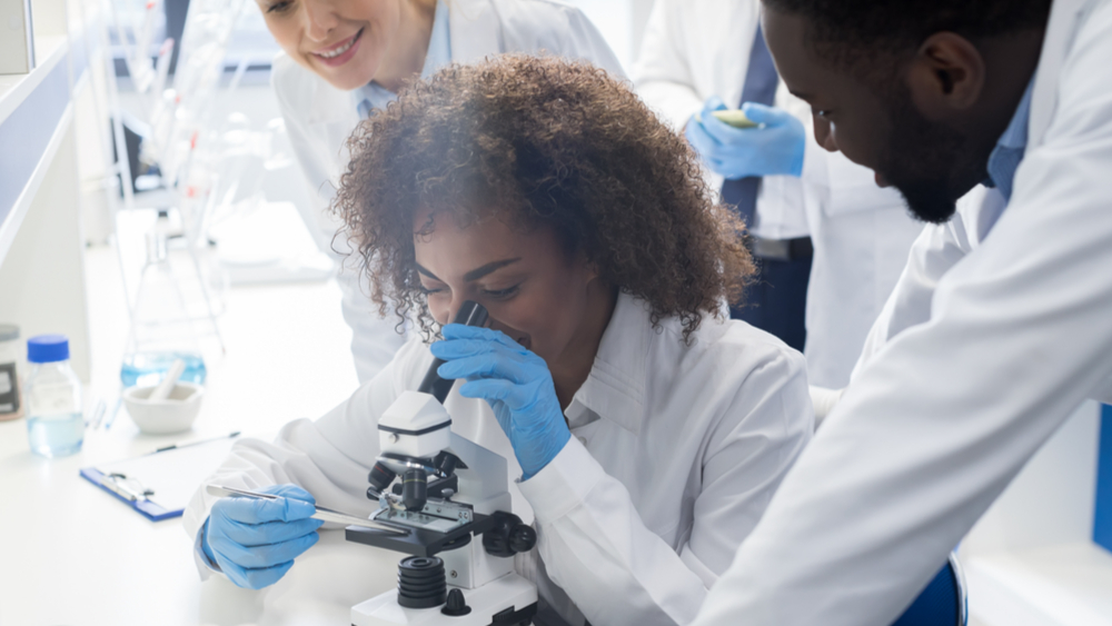 A group of 3 scientists gathered around a microscope, working together