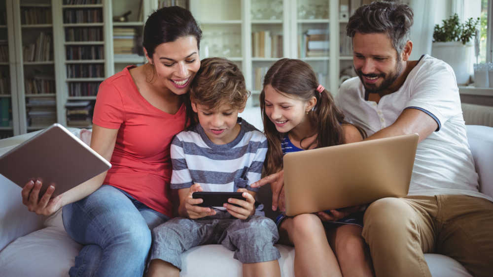 A family (two parents and two children) sit on the sofa using a laptop, tablet and smartphone together