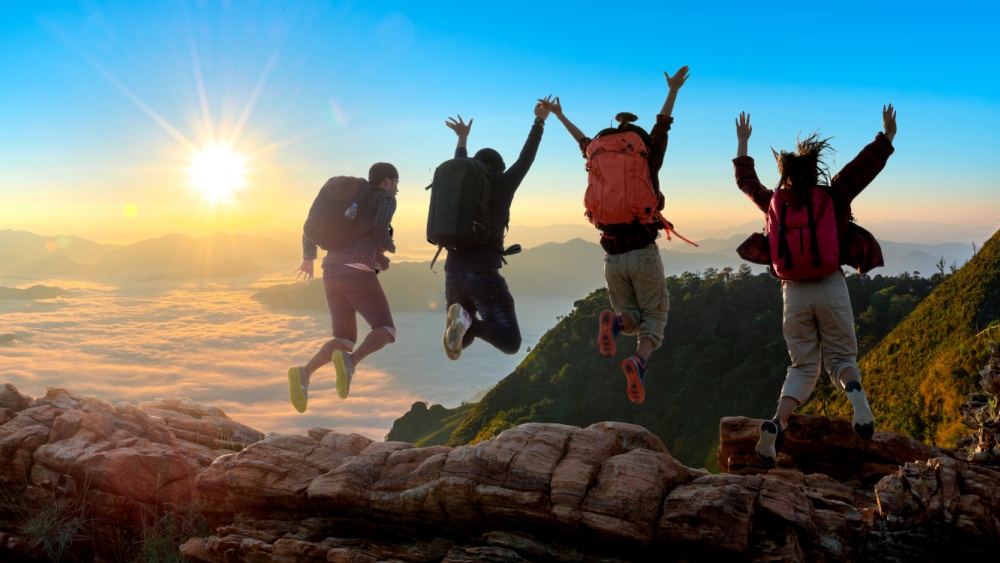 4 people are jumping on a mountain top with sun and clouds in the background