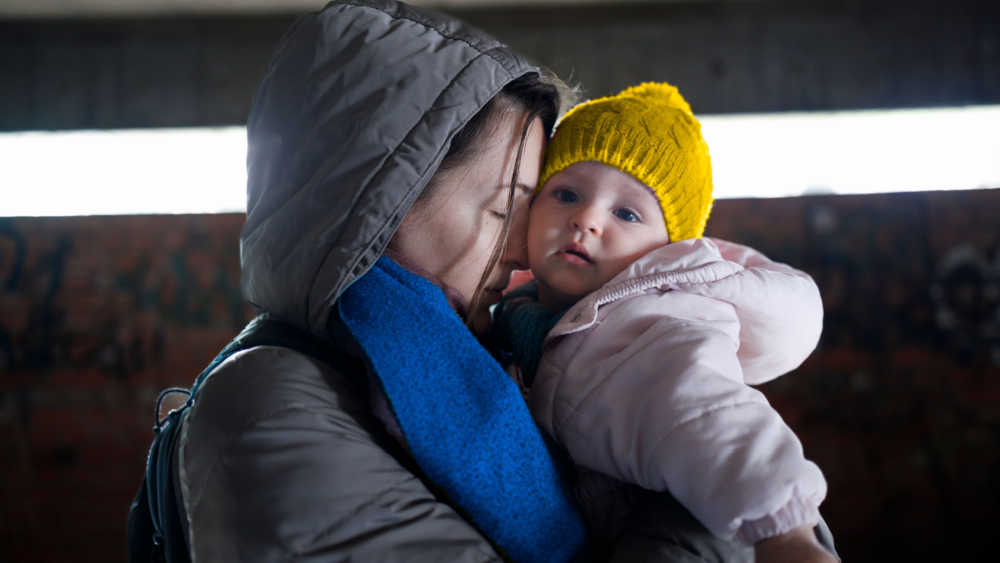 Women with blue wool scarf holds a baby with yellow beanie on her arms. Both dress in winter clothes.