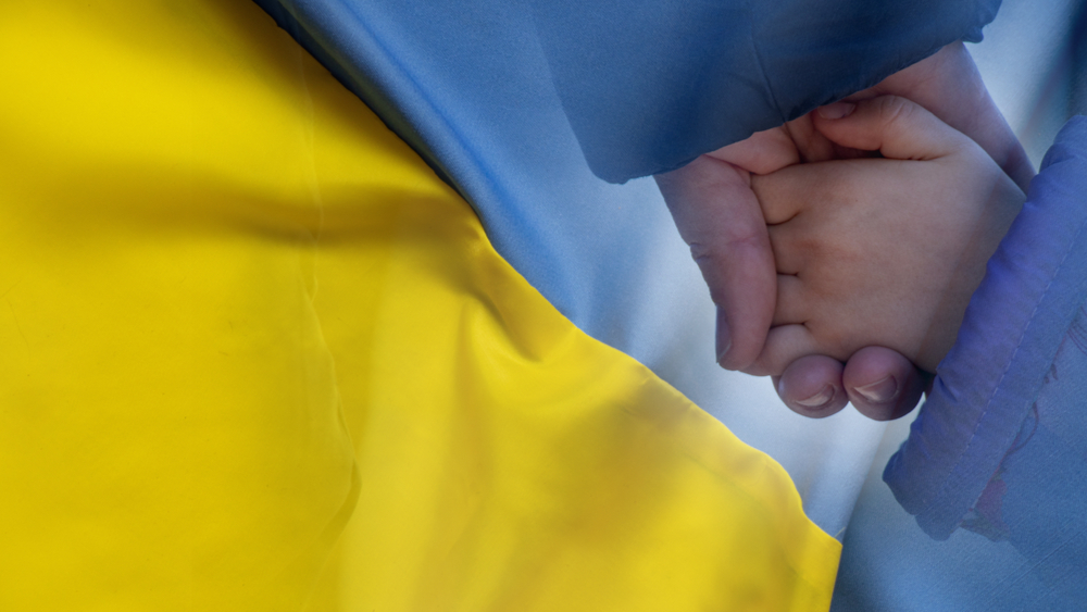 Adult and child hold hands with the Ukrainian flag in the background