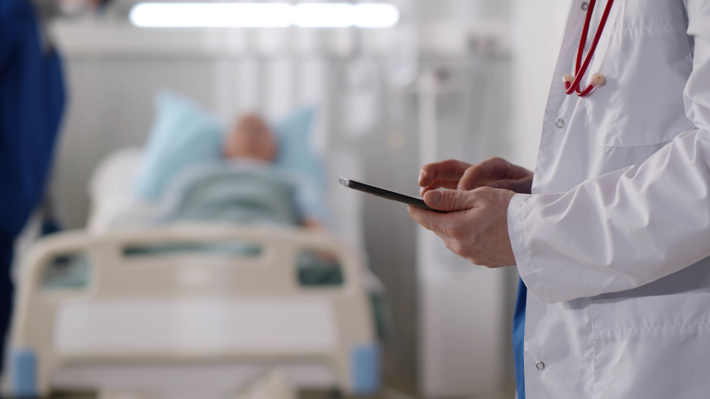 A man is lying in a hospital bed in the background. A nurse and a doctor is standing and the doctor types in some data on a handheld screen