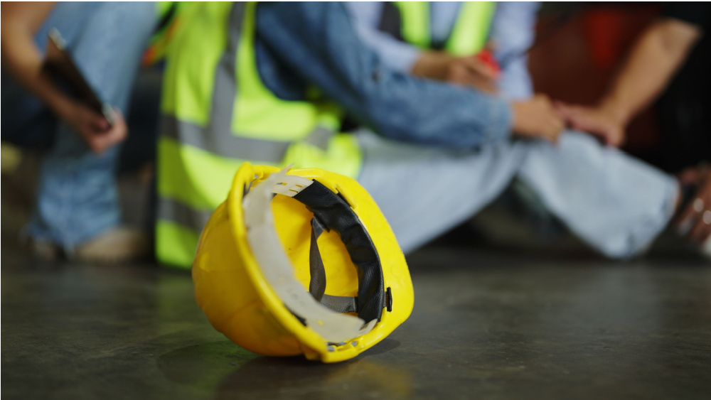 A person sitting on the ground dressed in the safety vest with a protective helmet next to him. People are gathered around him.