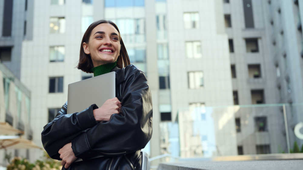 A person stands in an urban area smiling while hugging their laptop