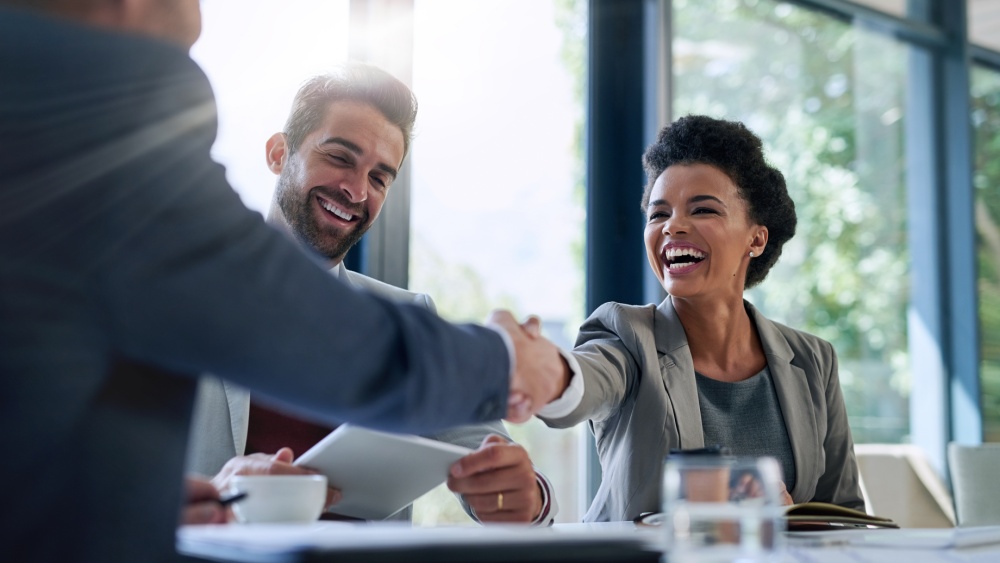 Three people sitting at the table in the business surrounding, two of them are shaking hands across the table.