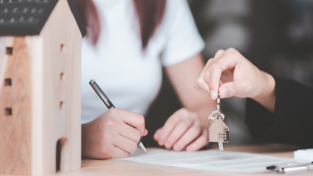 person holds the keys to something next to a mock up paper house, indicating it might be a house key and creating the idea that the other person is signing a contract to a house.