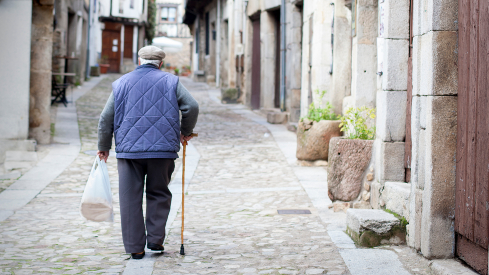 Elderly man with a cane walks down a quaint street while holding a shopping bag
