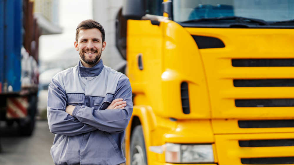 A happy truck driver is standing beside his yellow truck