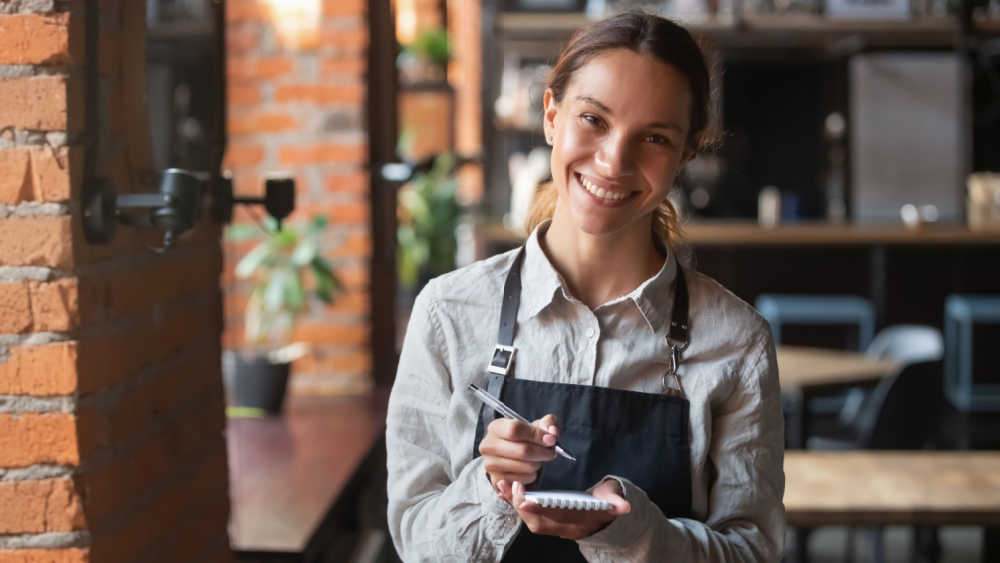 A waiter is standing and smiling in a restaurant and waiting to write down an order