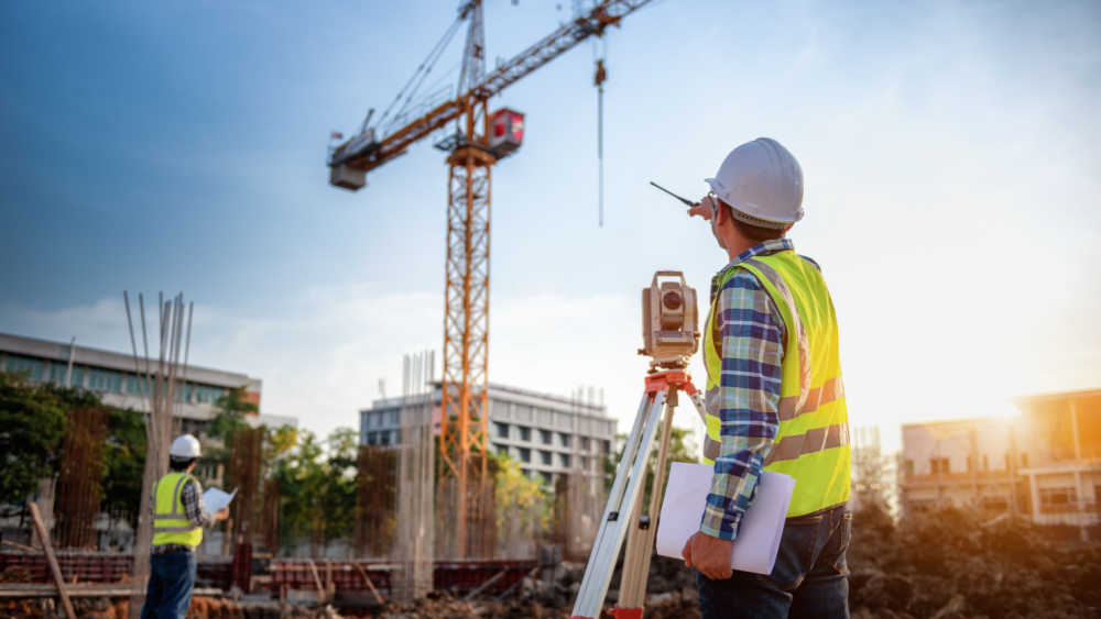 Two persons are standing on a construction site with a crane and some buildings in the background