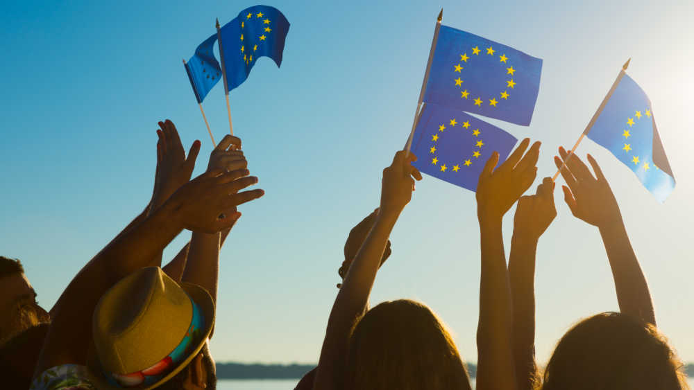 Hands waving EU flags towards the clear blue sky.