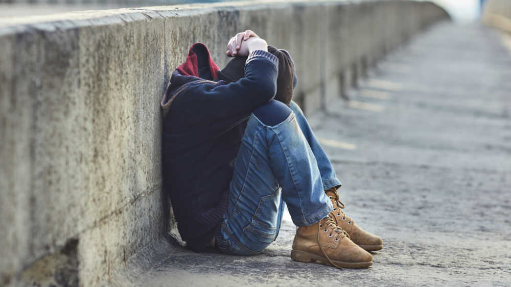 A young person is sitting on the pavement with his arms over his head hiding his face