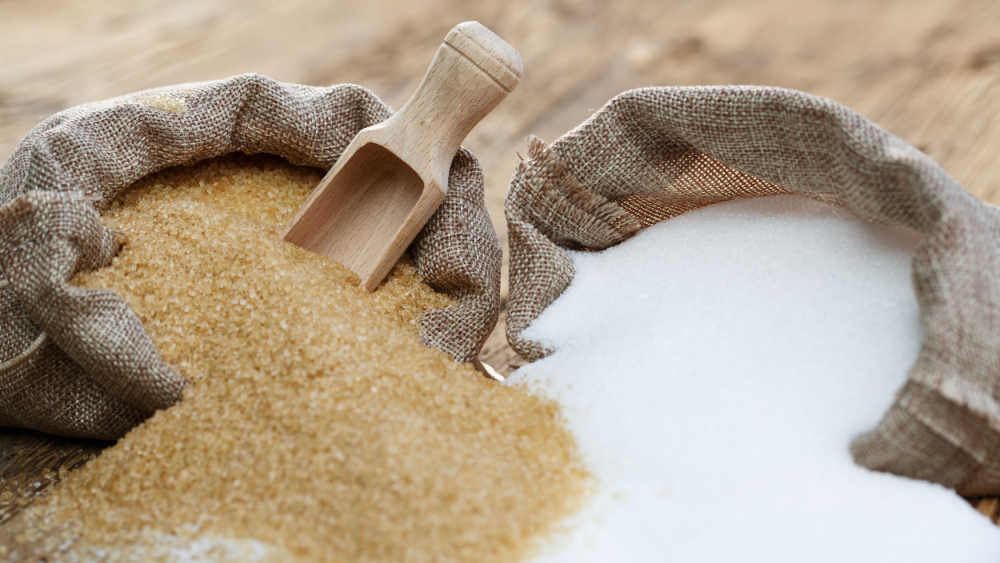 Two bags of sugar are standing on the ground. One bag is with brown sugar and one is with white sugar.