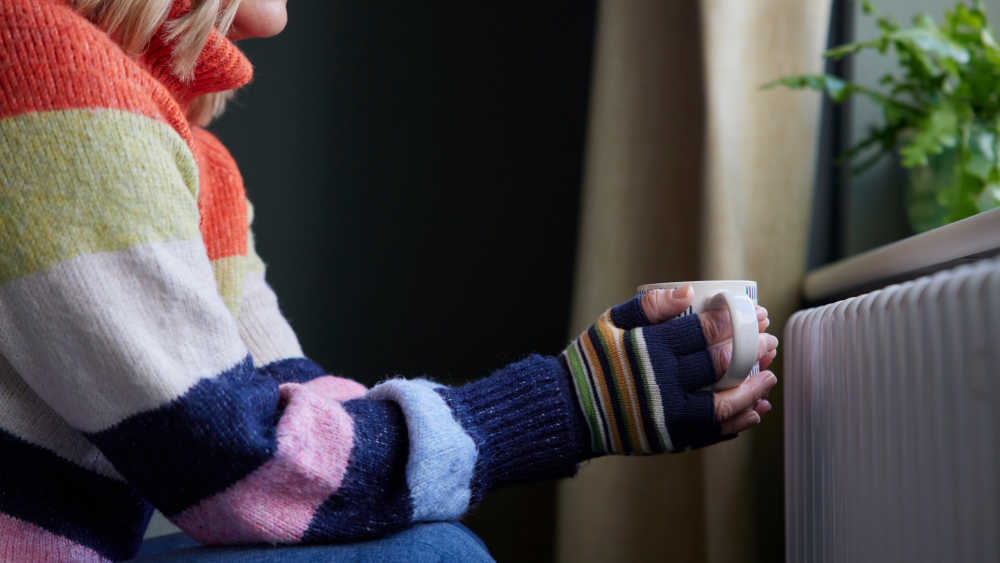 A person wearing a sweater and gloves, holding a mug beside a radiator.