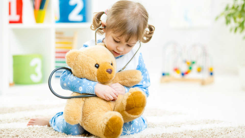 A child is sitting on a mattress in a room with toys holding her teddy