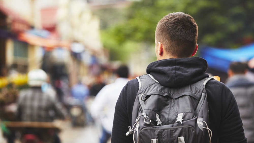 A young male person with a backpack is walking down a street with people sitting outside. Only the back of the person can be seen.