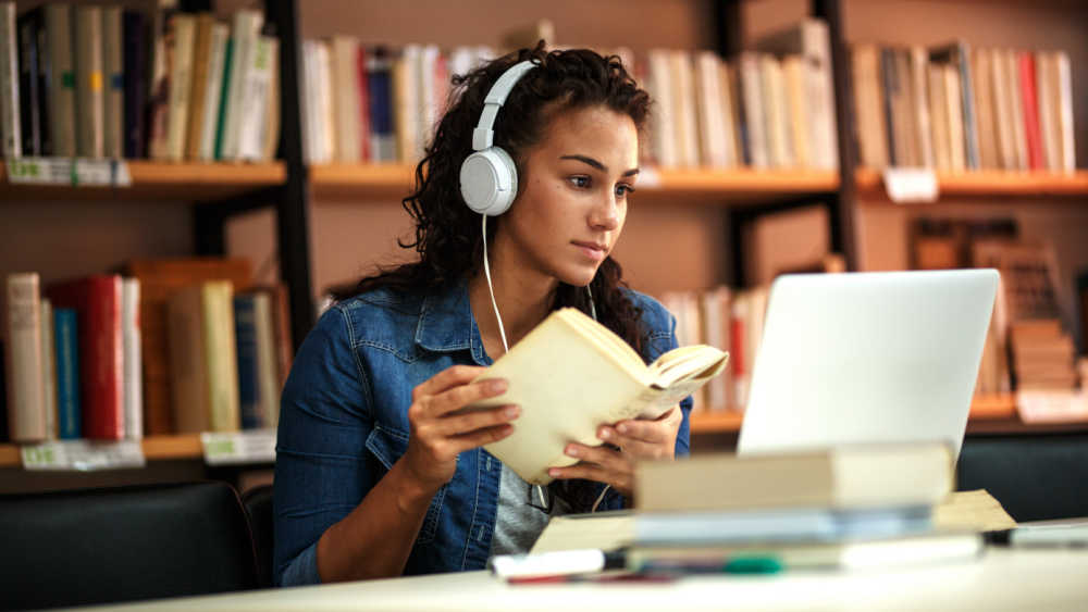 A young woman sits in a library, holding a book and wearing headphones while looking at her laptop