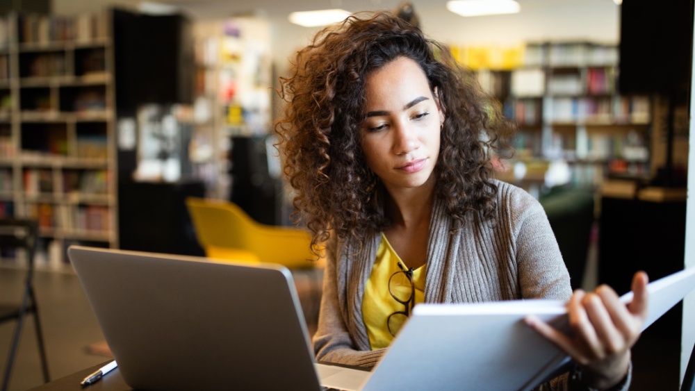 woman studying in a library with computer open and holding a book.