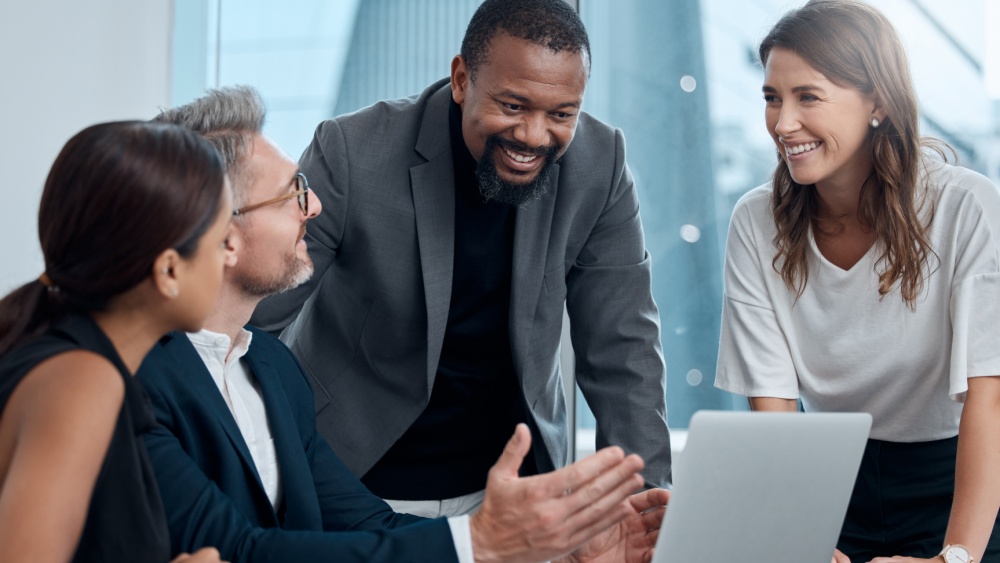 4 people standing and sitting in front of a laptop.