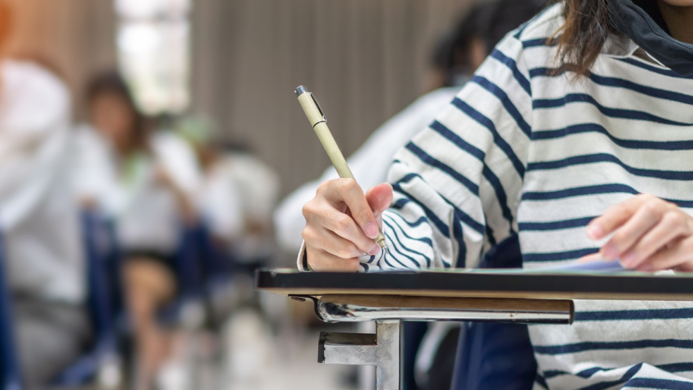 A person is sitting at a desk in a classroom with more people writing something on a paper