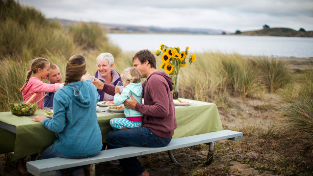 A group of people of different ages sitting around a table next to a lake sharing food.