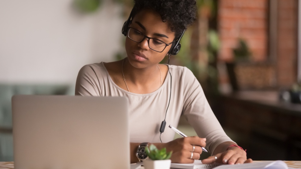 Woman wearing headphones with a pen in her hand while looking at a laptop.