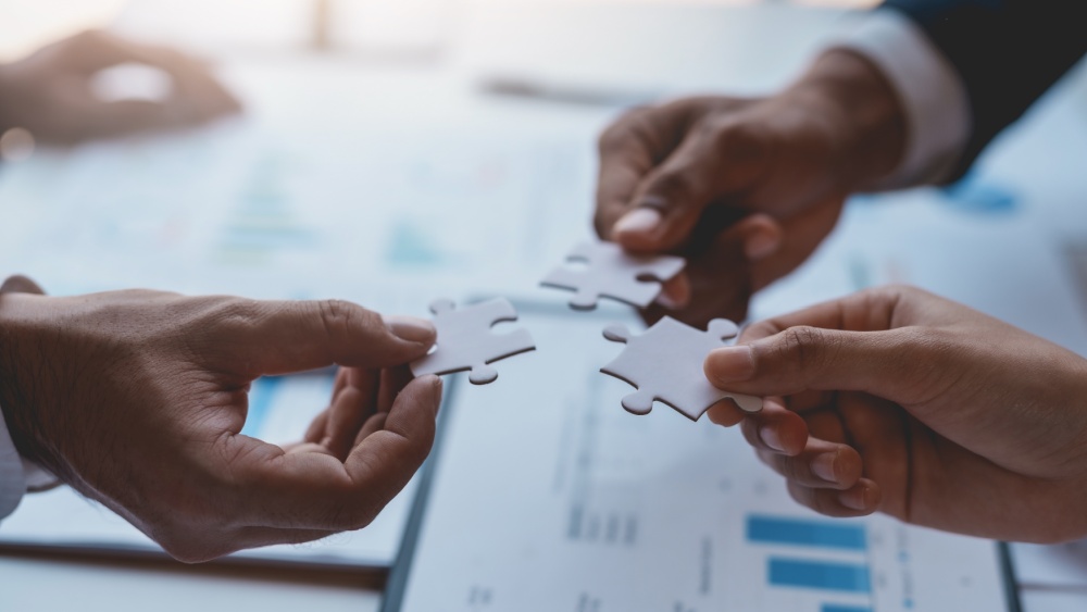 Three hands holding pieces of puzzles over the table containing documents with charts..