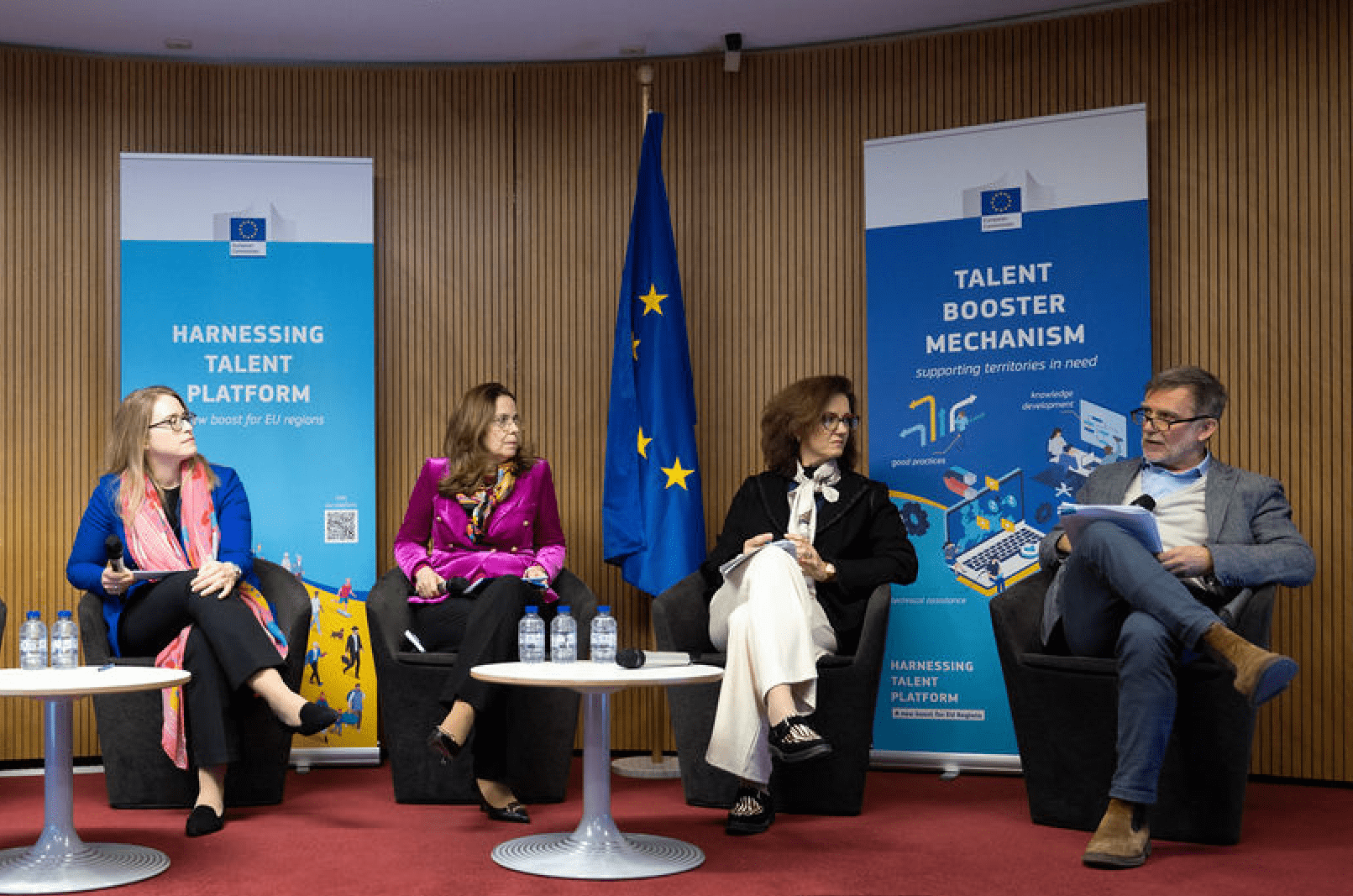 Four people sit on a panel stage with two EU flags and banners.