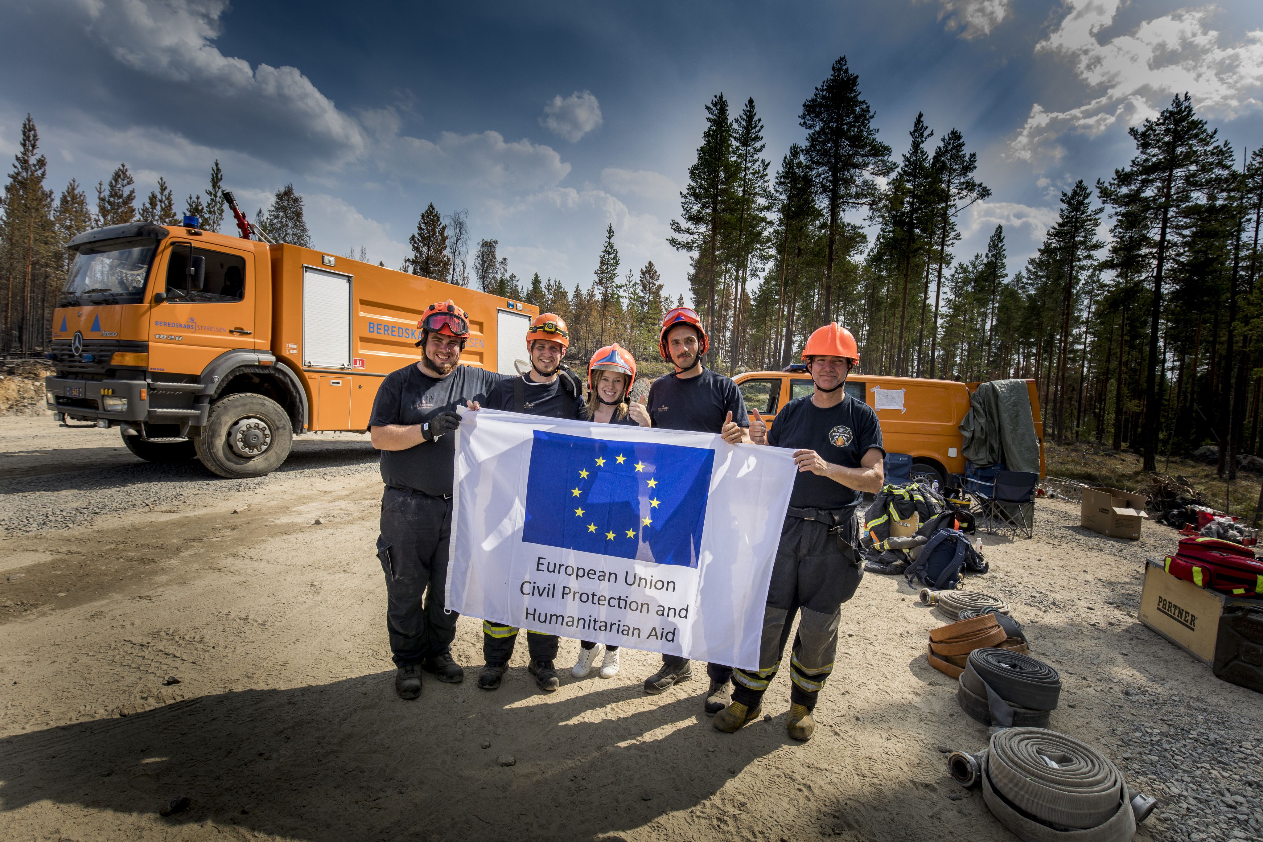 A group of firefighters hold up a European Union Civil Protection & Humanitarian Aid flag.